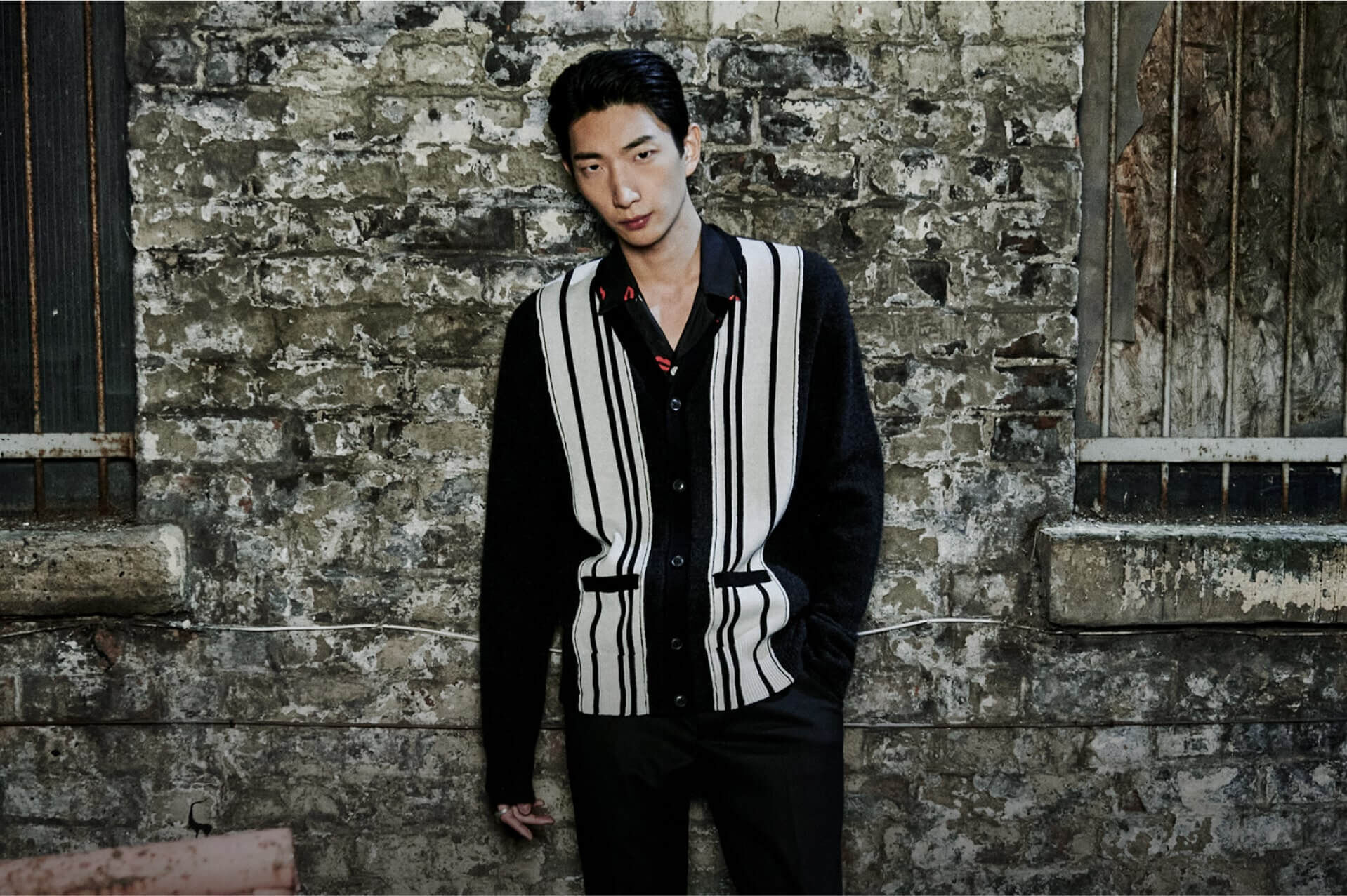 A man wearing a black and white striped cardigan leaning against a brick wall at night.