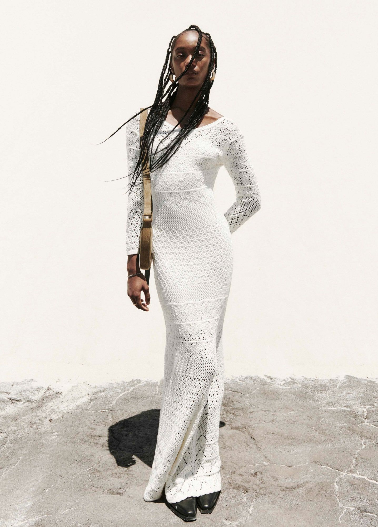 A model wearing white midi lace knit dress, large golden hoop earrings and a light brown suede crossbody bag standing in front of a beige background.