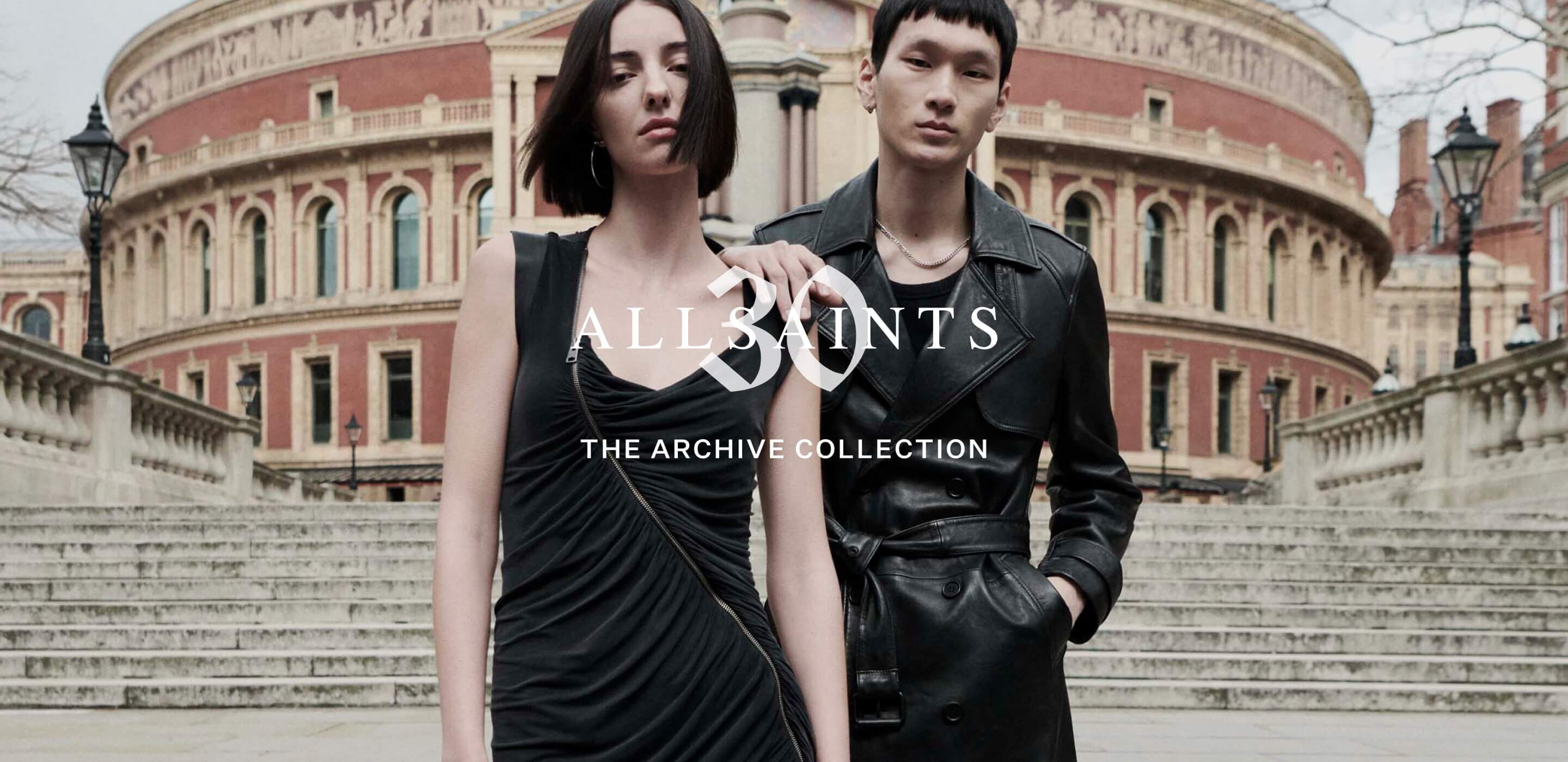 A male and female model standing in from of the Royal Albert Hall wearing a black asymmetrical dress and leather trench coat 