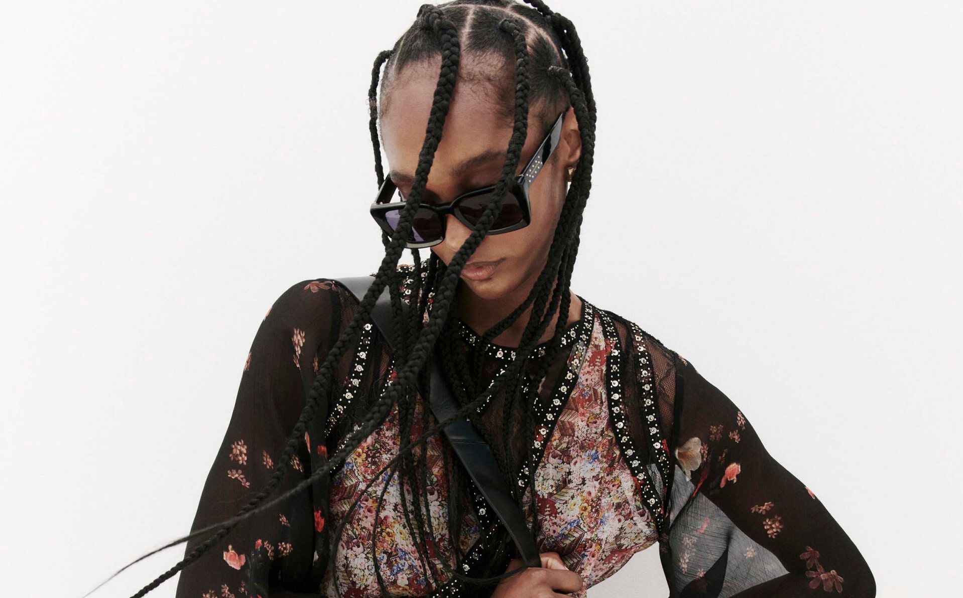 Closeup of a female model wearing a printed dress with panels and ornate trims and black sunglasses standing in front of a beige wall.