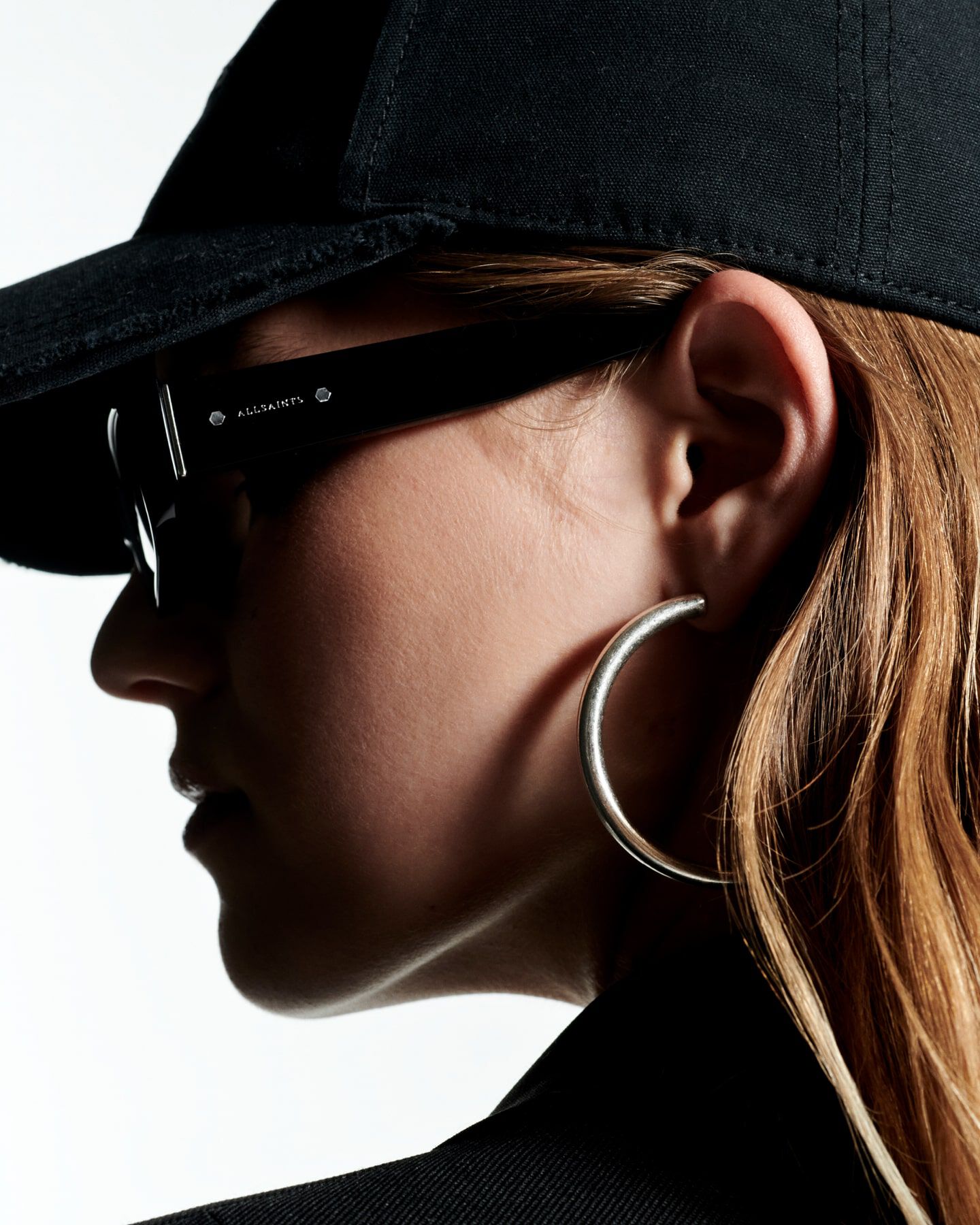 Closeup photograph of a female model wearing sunglasses, large hoop earrings and a baseball cap.