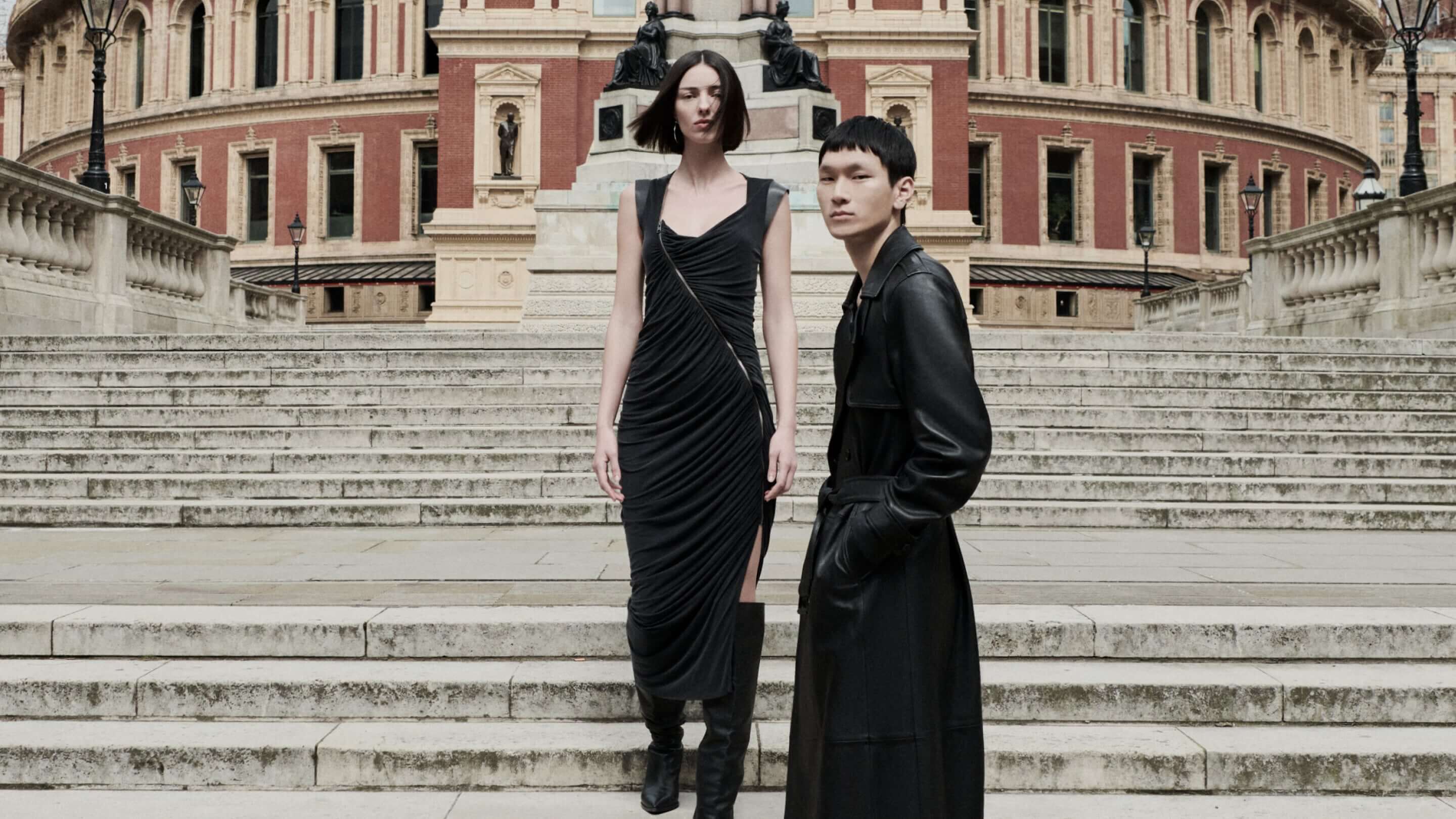 A male and female model standing in from of the Royal Albert Hall wearing a black asymmetrical dress and leather trench coat 