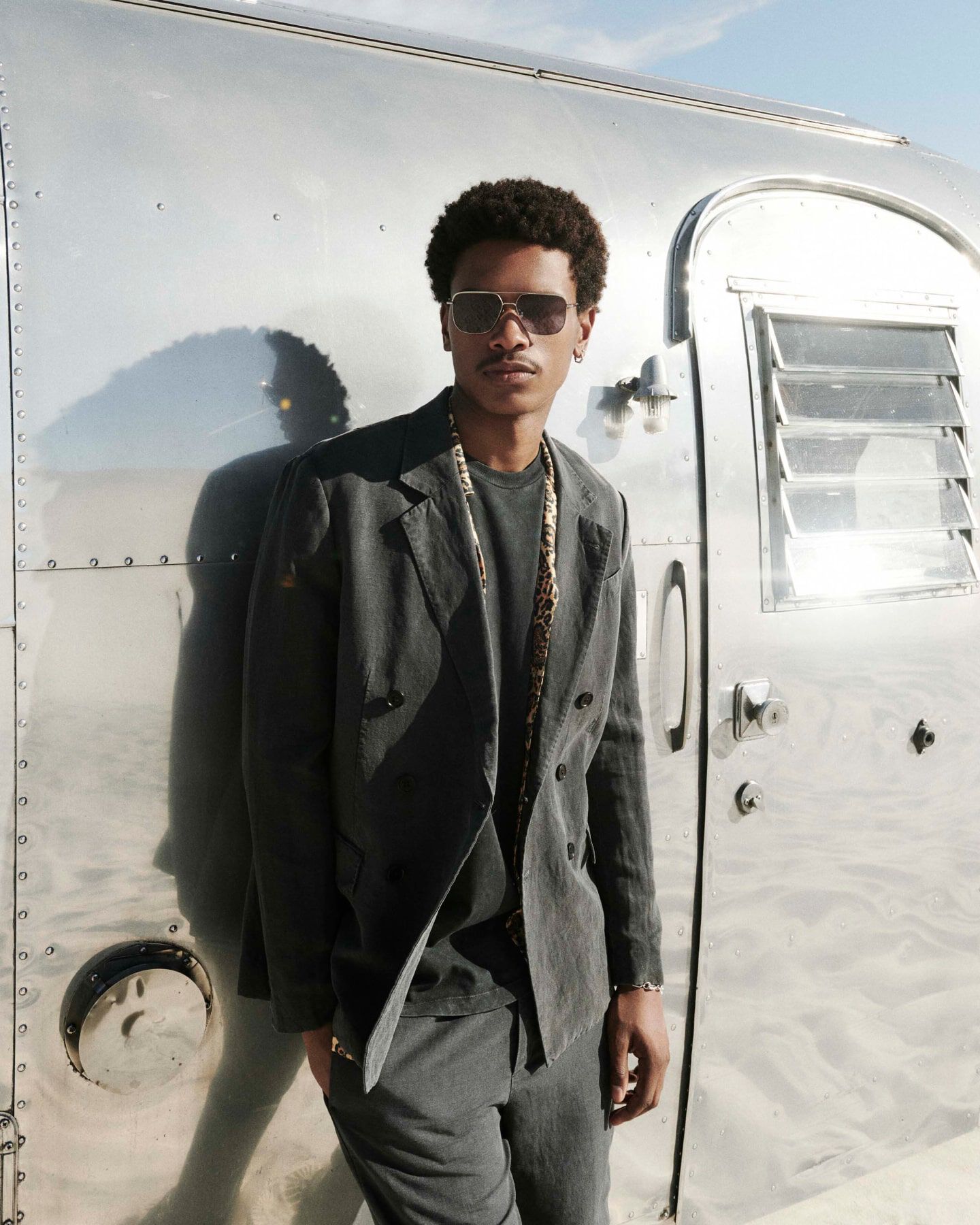 Man leaning against an airplane, wearing sunglasses, a grey suit and leopard print shirt over a black t-shirt.