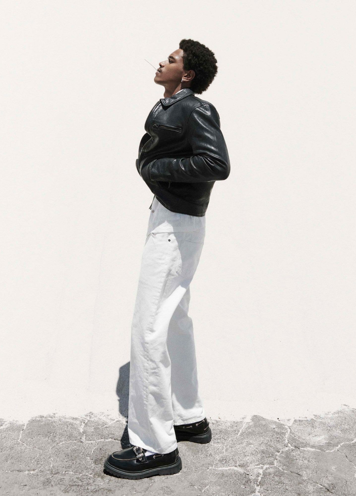 A man wearing a black leather jacket, white pants and black leather loafers standing in front of a white wall.