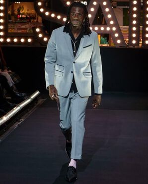 A man wearing a grey suit with a black shirt and dress shoes catwalking.