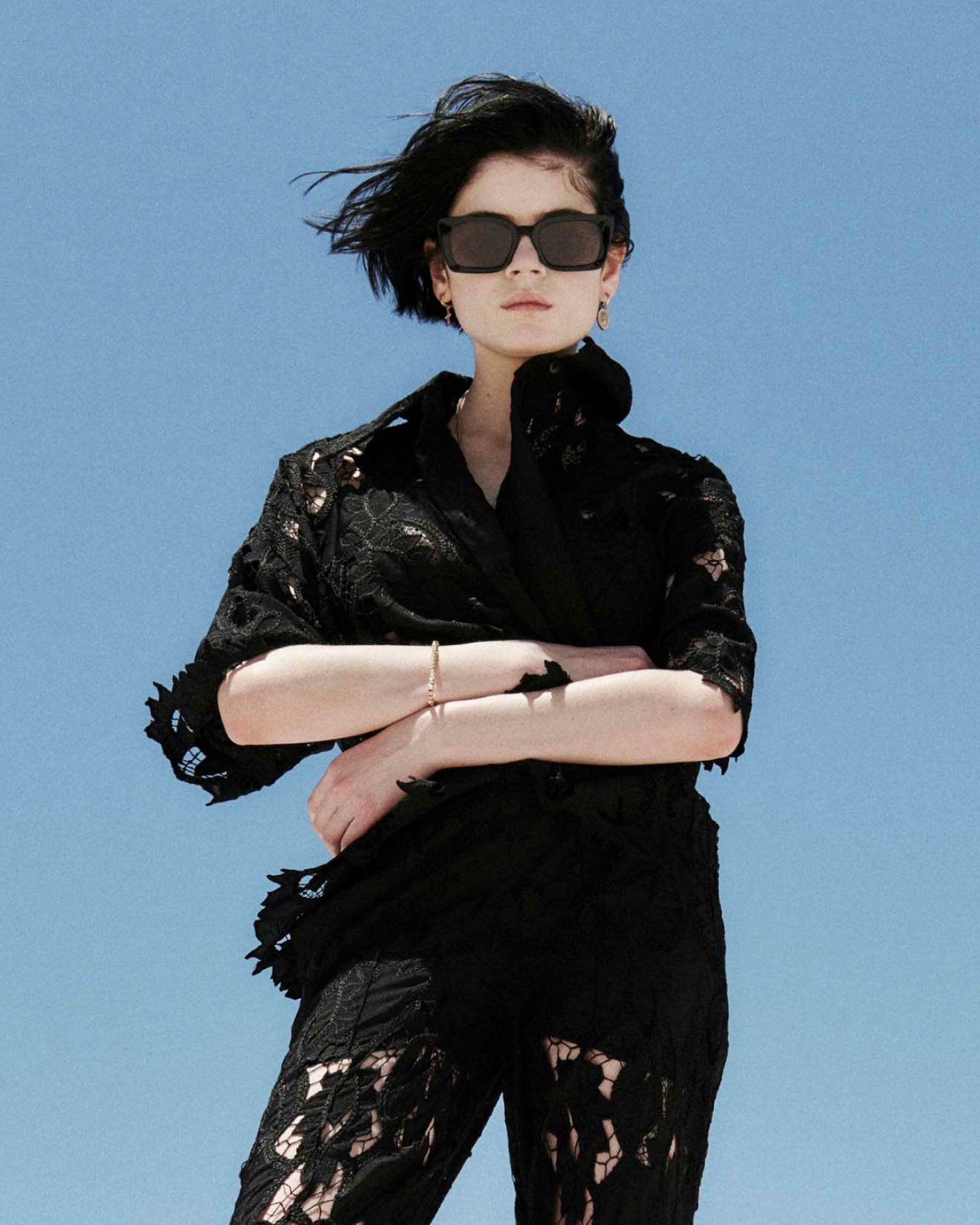 Female model wearing black sunglasses and a black shirt and pants in a floral lace cutout fabric standing in front of a blue sky.