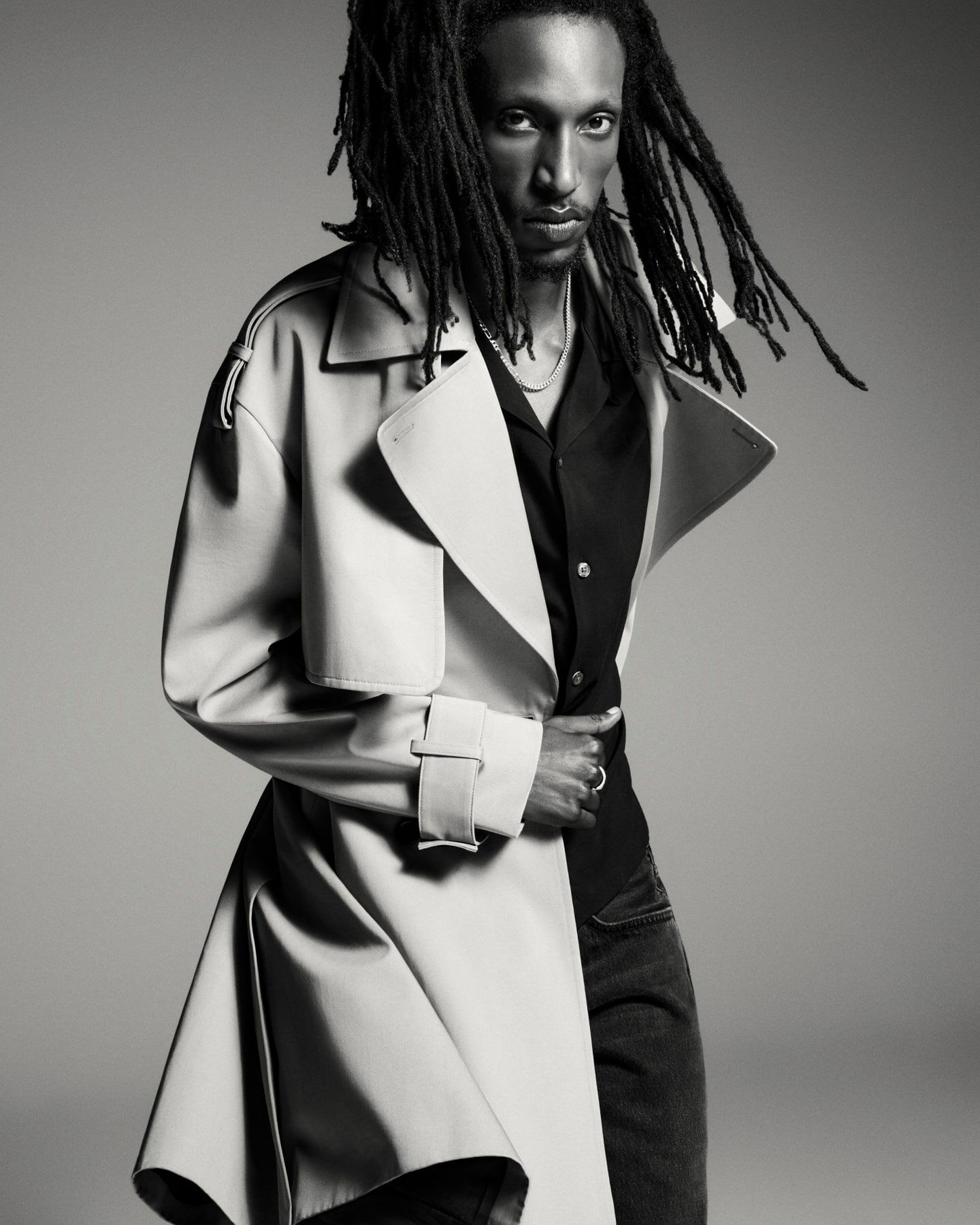 A black and white photograph of a man wearing a trench coat over a black shirt.