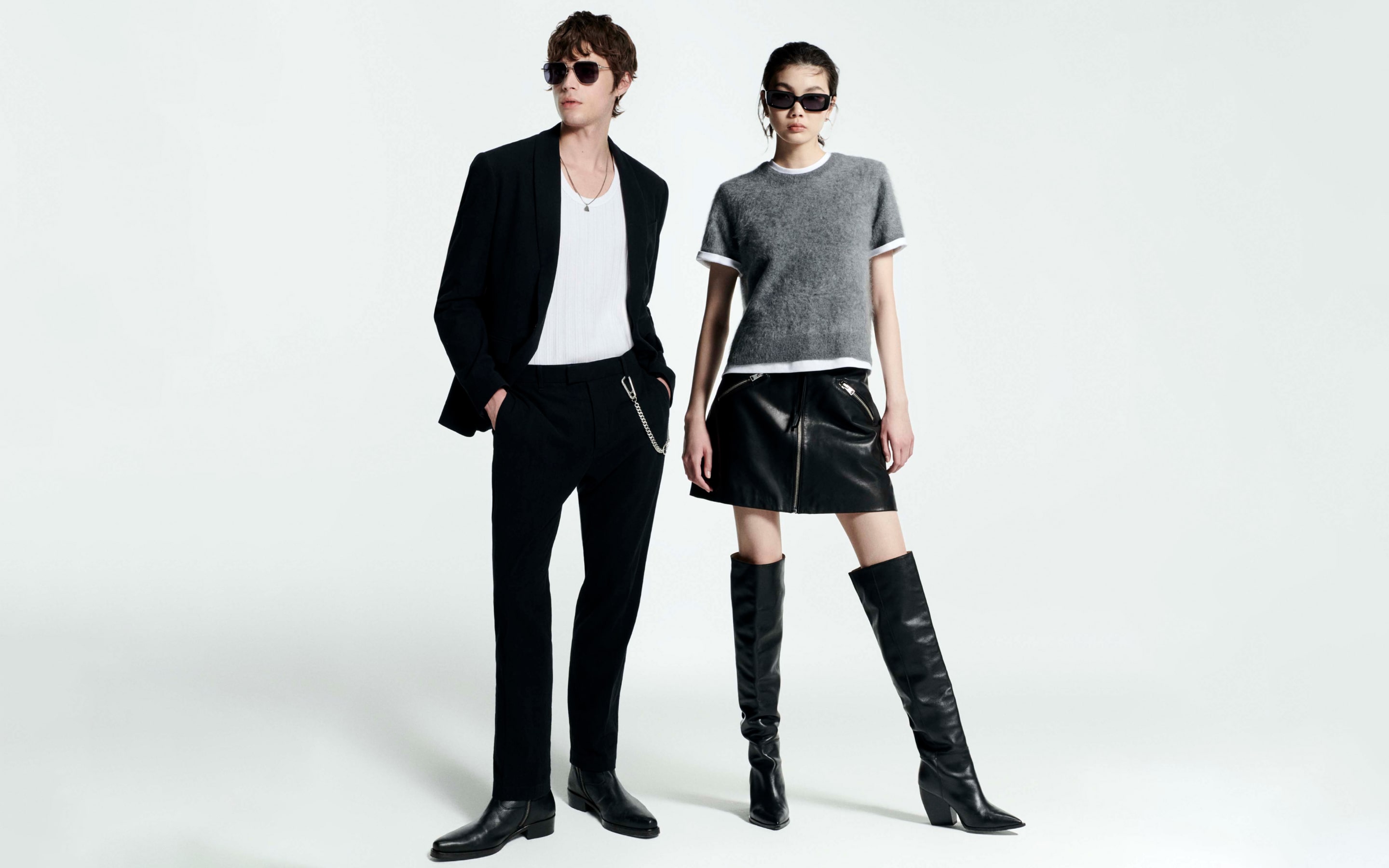 A man wearing black sunglasses, a black blazer, black trousers and a white vest standing next to a woman with a grey brushed jumper, a black leather mini skirt and black thigh high leather boots.